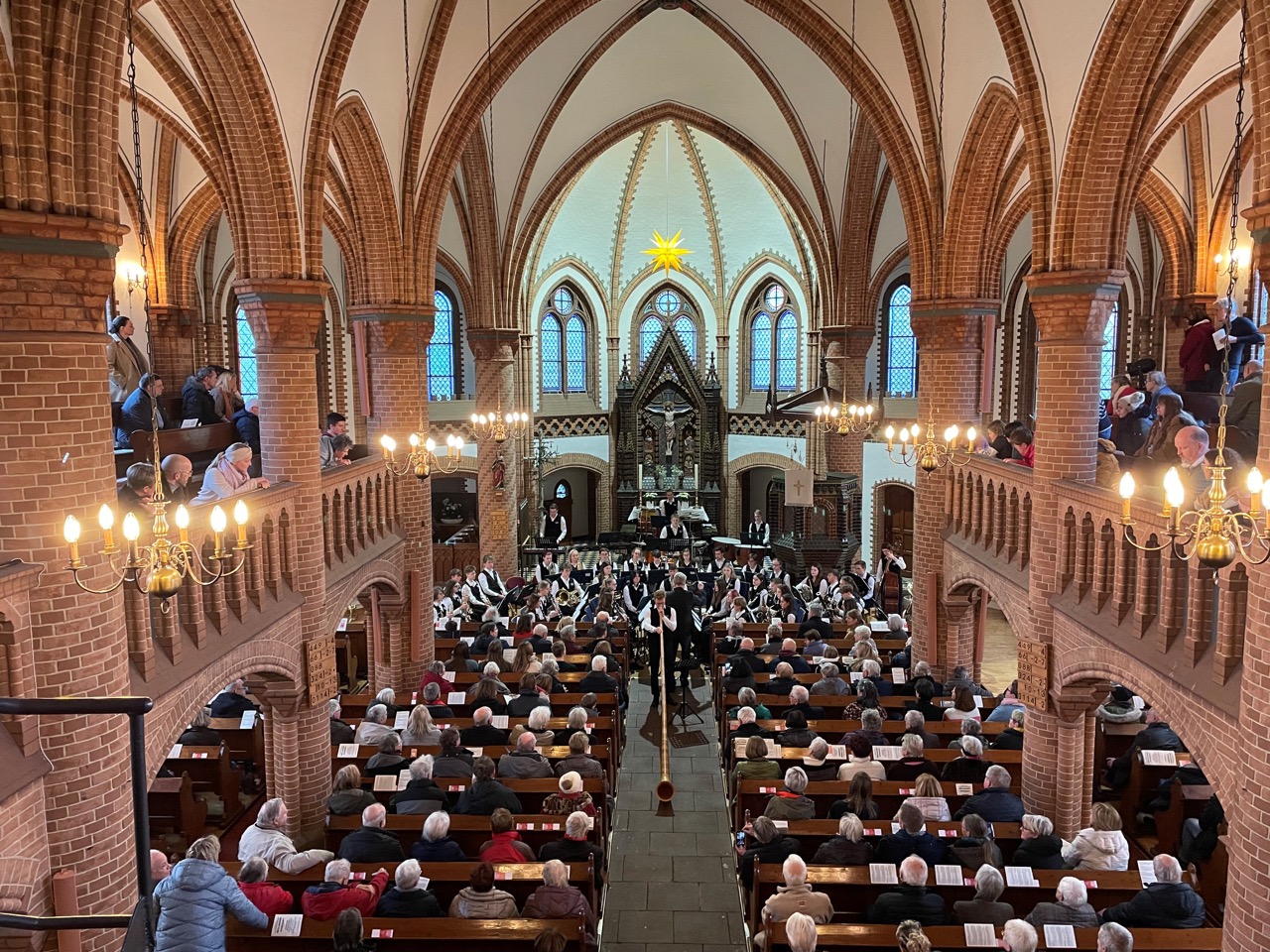 Die Ratsband Stadthagen gibt ein Konzert in der warm beleuchteten Kirche während ihres Neujahrskonzerts 2024 in Hagenburg. Der Dirigent steht zentral vor dem Orchester, und die Zuschauer füllen sowohl das Erdgeschoss als auch die Empore der Kirche. Die gotische Architektur, die hohen Gewölbedecken und die leuchtenden Kronleuchter bieten eine majestätische Kulisse für die Veranstaltung.